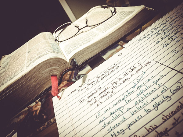 An open Bible with reading glasses and handwritten sermon notes on a desk, representing the study and craft behind Christian writing and self-publishing